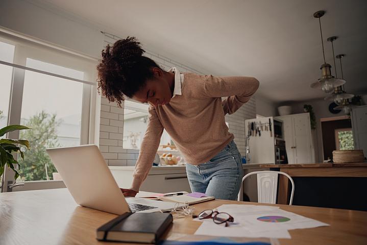 Young woman standing and holding her back while working on a laptop at home Young woman standing and holding her back while working on a laptop at home