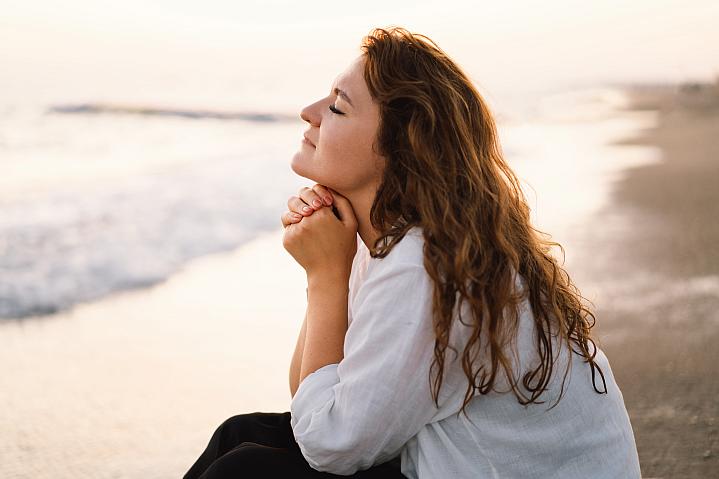 Woman with her eyes closed and hands clasped sitting next to the sea.